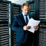 A professional male executive, mid-30s, in a modest business suit, stands in a cluttered, dimly lit server room filled with bulky, outdated server racks. He holds a stack of physical documents, appearing stressed and overwhelmed by the sheer volume. The environment emphasizes limited capacity and manual data handling. Perfect anatomy, correct proportions, natural pose, well-formed hands, proper finger count, natural body proportions, fully clothed, appropriate attire, safe for work, professional, appropriate content, family-friendly, high-quality, professional photography.
